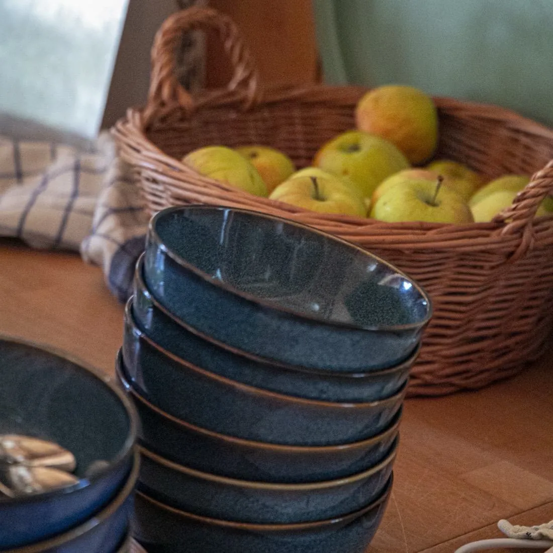Gestapelte blaue Keramikschalen vor einem Weidenkorb mit Äpfeln auf dem Holztisch
